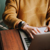 Eine Person sitzt an einem Tisch vor einem aufgeklappten Laptop. Abgebildet ist ein Ausschnitt des Oberkörpers in einem gelben Pullover. Die Hände tippen auf der Tastatur.