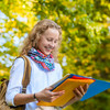 Standing female student in front of green foliage holds a few binders in her hand and directs her gaze towards them