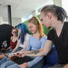 Three teenagers are sitting on a sofa and working on two laptops. A fourth person in the foreground is explaining something to one of the teenagers.