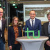 A woman and three men stand in a row. In front of them is the TU Dortmund logo on a high table, behind them is a presentation on a screen.