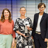 Three women pose side by side for a photo. The woman in the middle is holding an award in her hand.