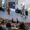 Three people stand in the packed Audimax during the DLR_Space_Show. They wear costumes of an earth, a kangaroo and an astronaut. The astronaut jumps into the air.