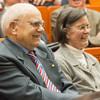 An elderly man and woman sit in a lecture hall and look ahead smiling