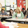 Pupils sit in a class and write in notebooks sitting in front of them a teacher at a desk.