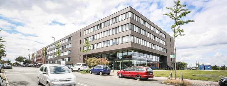 ITMC: Ein großes, braun-rot verklinkertes Gebäude mit Flachdach aus der Froschperspektive fotografiert. Davor eine Straße, auf der ein Auto fährt. Andere Autos stehen am Straßenrand hintereinander geparkt.