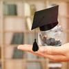 A transparent piggy bank full of coins and with a doctoral cap is held in front of a book shelf. 